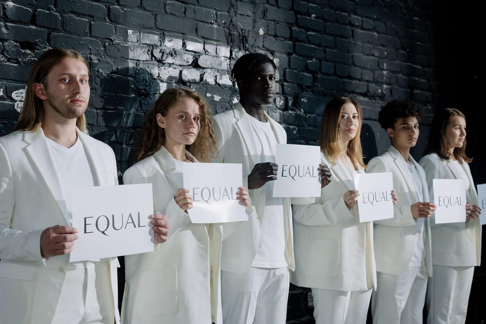 Six people dressed identically in white suits each holding a piece of paper with word 'equal' printed on it.